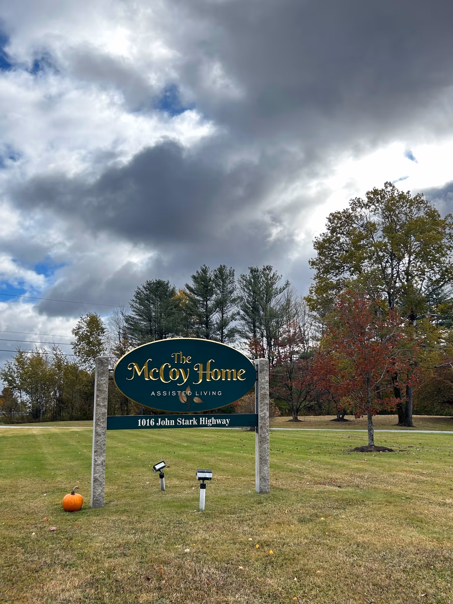 Outdoor view of a green and gold sign for The McCoy Home Assisted Living located at 1016 John Stark Highway, set on a grassy area with trees in the background under a cloudy sky. A small pumpkin is placed on the grass near the sign.