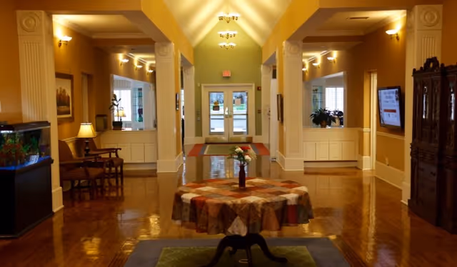 Sunlit lobby with a central round table, polished wood floors, columns, seating areas and double glass entrance doors.