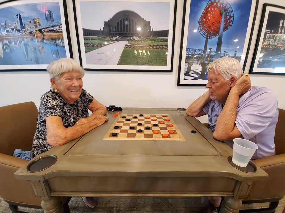 Two elderly people sitting at a table playing a game of checkers. The woman on the left is smiling and the man on the right is resting his head on his hand, concentrating on the game. Behind them are framed photos of cityscapes and landmarks on the wall.