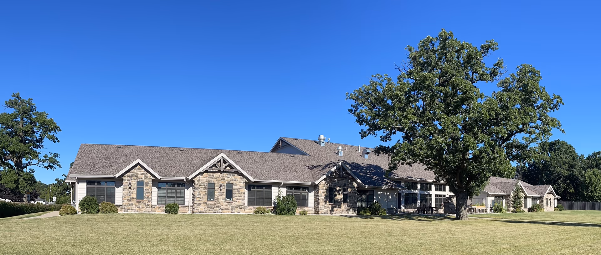 Exterior view of a single-story senior living facility building with stone and siding facade, multiple windows, and a large tree on a well-maintained grassy lawn under a clear blue sky.