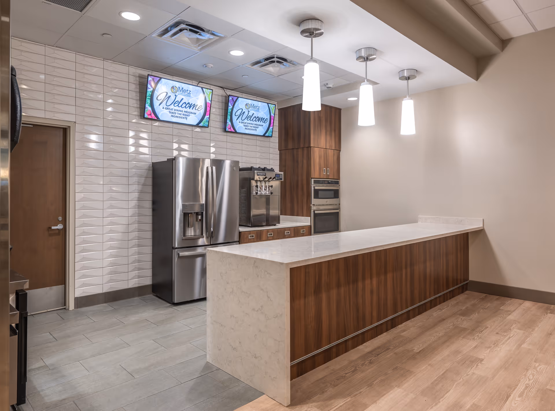 Modern kitchen area with a large marble countertop island, wooden cabinetry, stainless steel refrigerator, built-in oven, and two digital screens on the tiled wall displaying a welcome message.