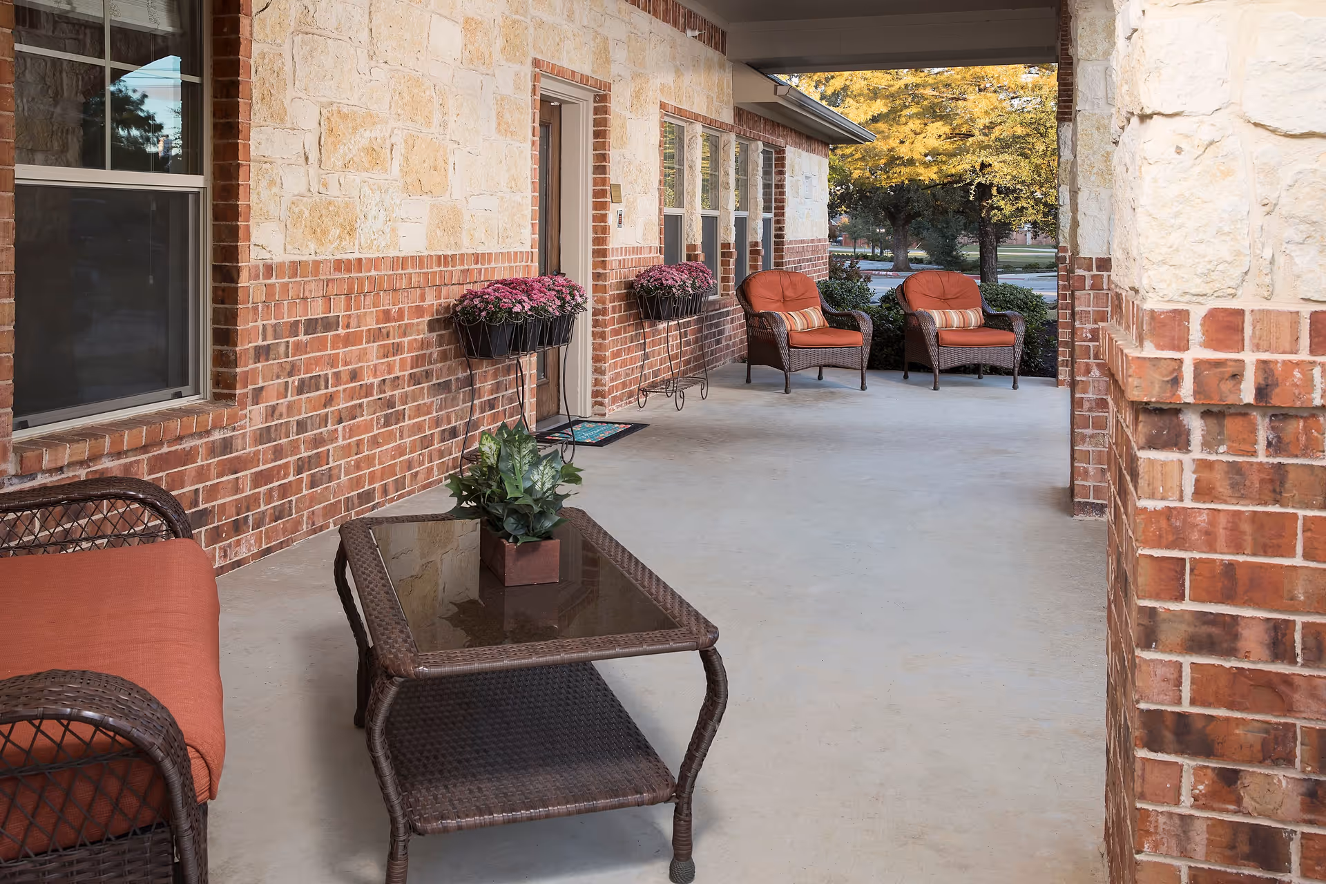 Covered outdoor patio area with brick and stone walls, featuring wicker furniture with orange cushions, a glass-top coffee table with a potted plant, and flower pots mounted on the wall. Trees and a street are visible in the background.