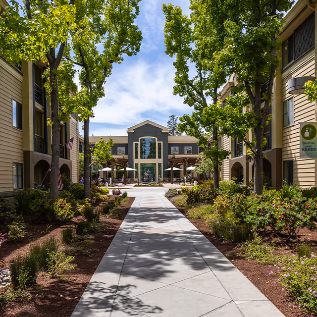 A sunny outdoor courtyard at The Terraces of Los Gatos featuring a wide concrete walkway flanked by landscaped garden beds with green trees and shrubs. Two three-story beige buildings with balconies and windows line both sides of the walkway, leading to a central building with large windows and outdoor seating under umbrellas. A sign on the right side reminds visitors to sanitize their hands.