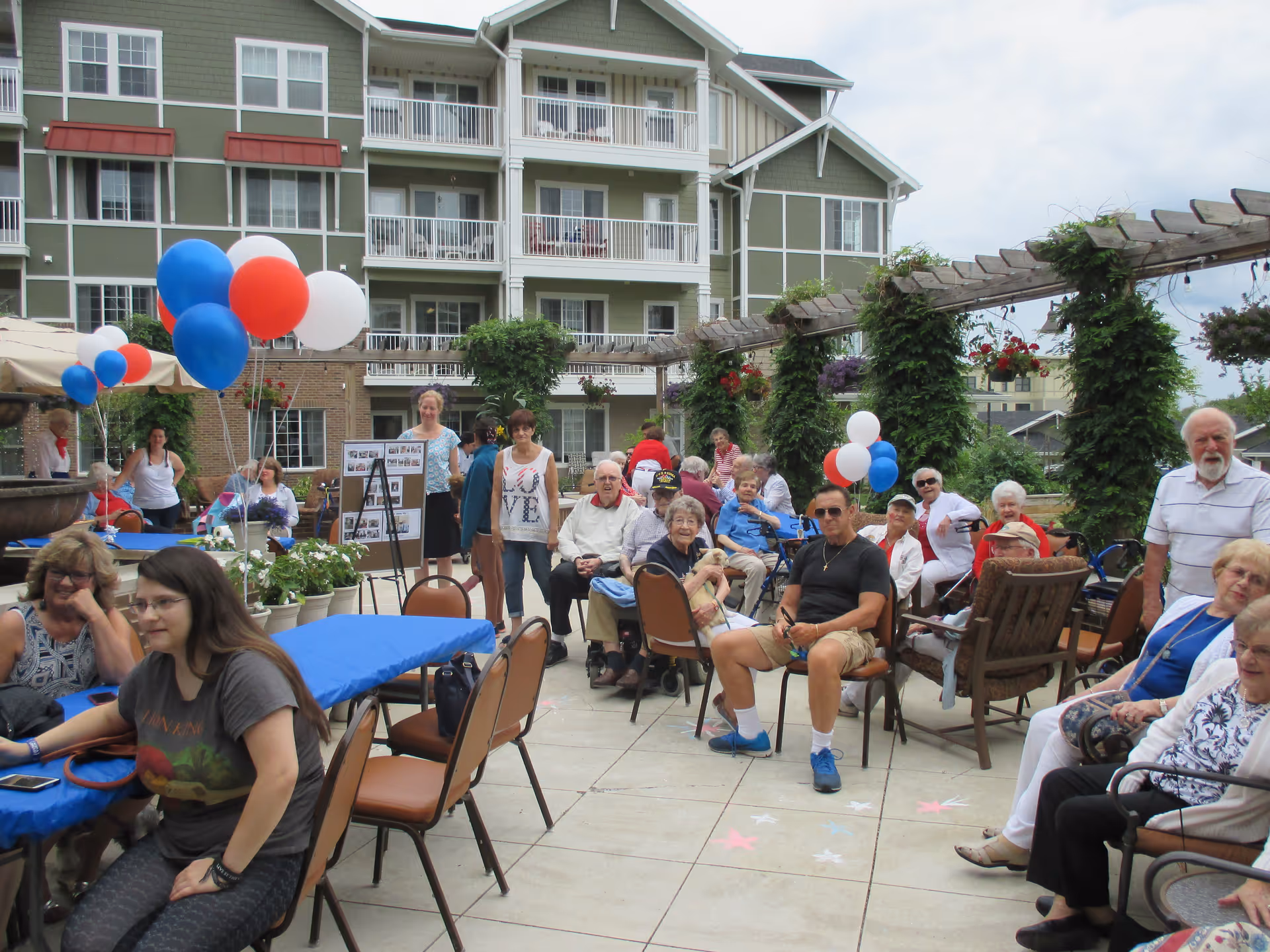 Group of senior residents and visitors seated at tables in an outdoor courtyard of a retirement community decorated with red, white, and blue balloons.