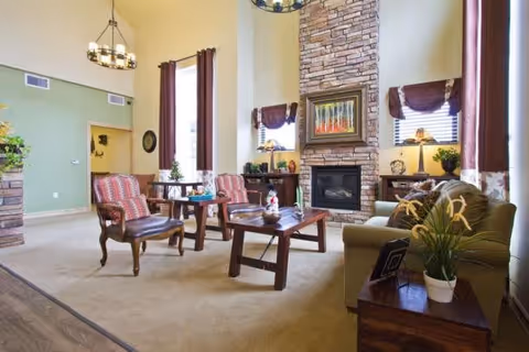 Spacious senior living lounge with armchairs, a sofa, coffee tables, and a stone fireplace under tall windows.