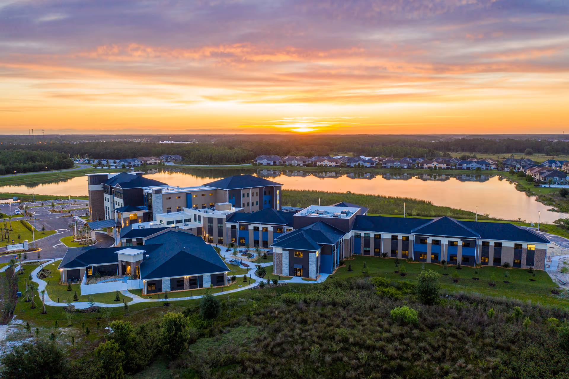 Aerial view of Blue Heron Senior Living facility at sunset, showing a large modern building complex with multiple wings, surrounded by landscaped grounds and a lake in the background reflecting the colorful sky.