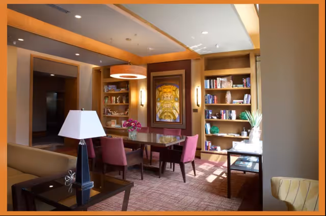 A cozy interior room with a rectangular wooden table surrounded by six pink upholstered chairs. Behind the table are built-in wooden bookshelves filled with books and decorative items. A large framed artwork hangs on the wall between the bookshelves. The room is warmly lit with ceiling lights and wall sconces, and there is a table lamp on a side table in the foreground. Sunlight streams in from the right side, illuminating part of the carpeted floor.