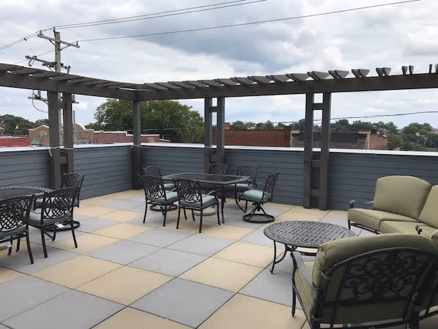 Outdoor patio area with metal tables and chairs, cushioned armchairs, and a pergola overhead. The patio is surrounded by a gray fence with a view of nearby buildings and trees under a cloudy sky.