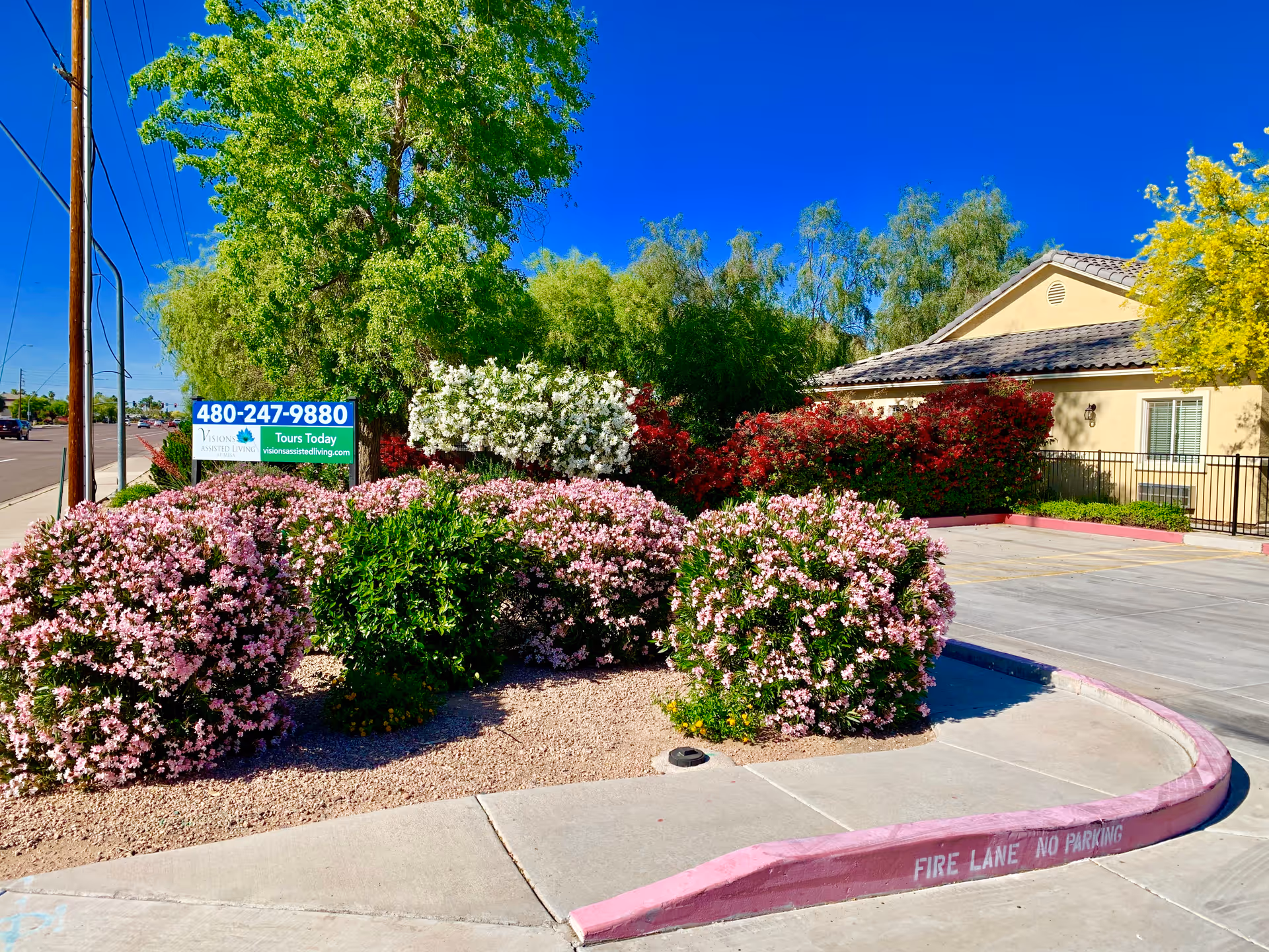 Front exterior of Visions Assisted Living showing flowering shrubs, a sign with a phone number, and the facility building under a clear blue sky.