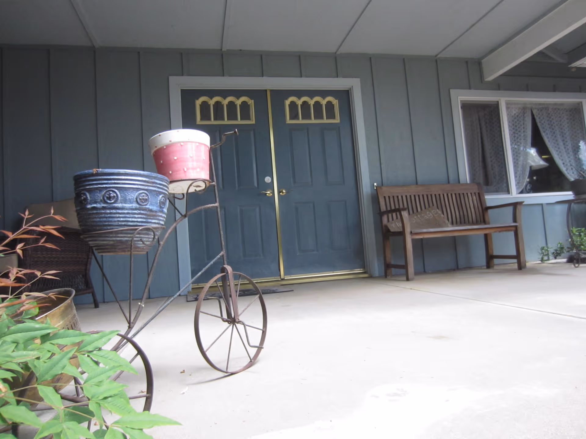 Covered porch with double blue doors, a wooden bench, and decorative bicycle planters in front.