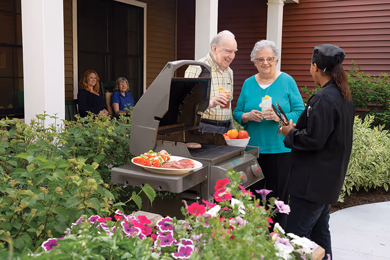 An outdoor barbecue scene at a senior living facility with a man and woman holding drinks and talking to a chef standing next to a grill with food. Two other women are seated on a porch in the background, surrounded by greenery and colorful flowers.