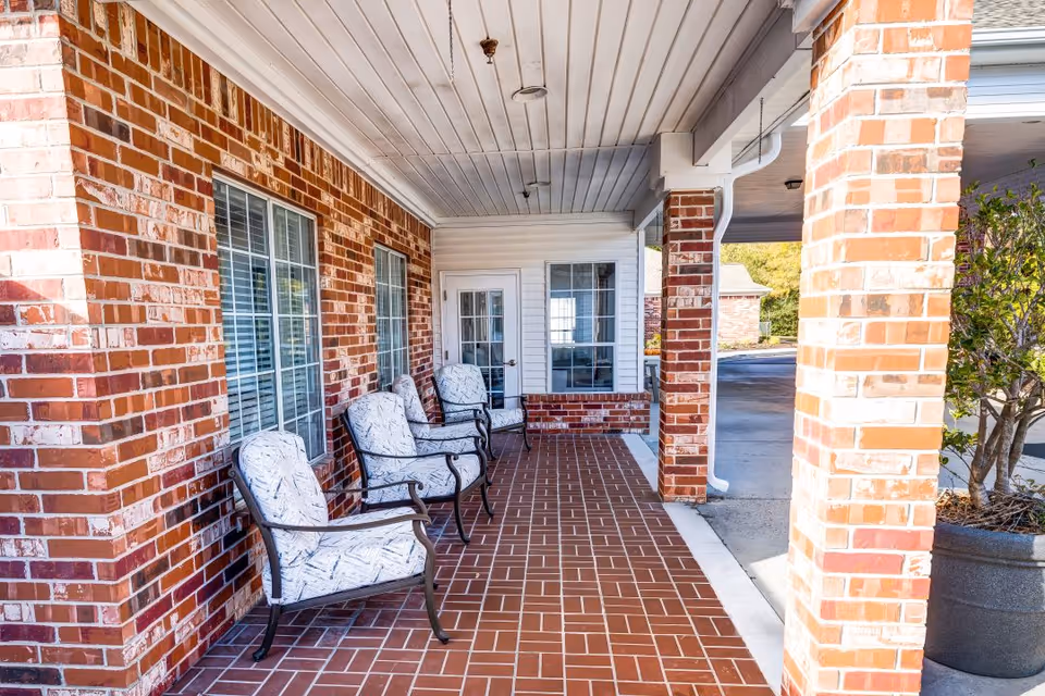 Covered outdoor patio area with brick walls and floor, featuring four cushioned chairs lined up against the wall. There are windows and a door with glass panes on the brick wall, and a potted plant is visible on the right side near a brick column.