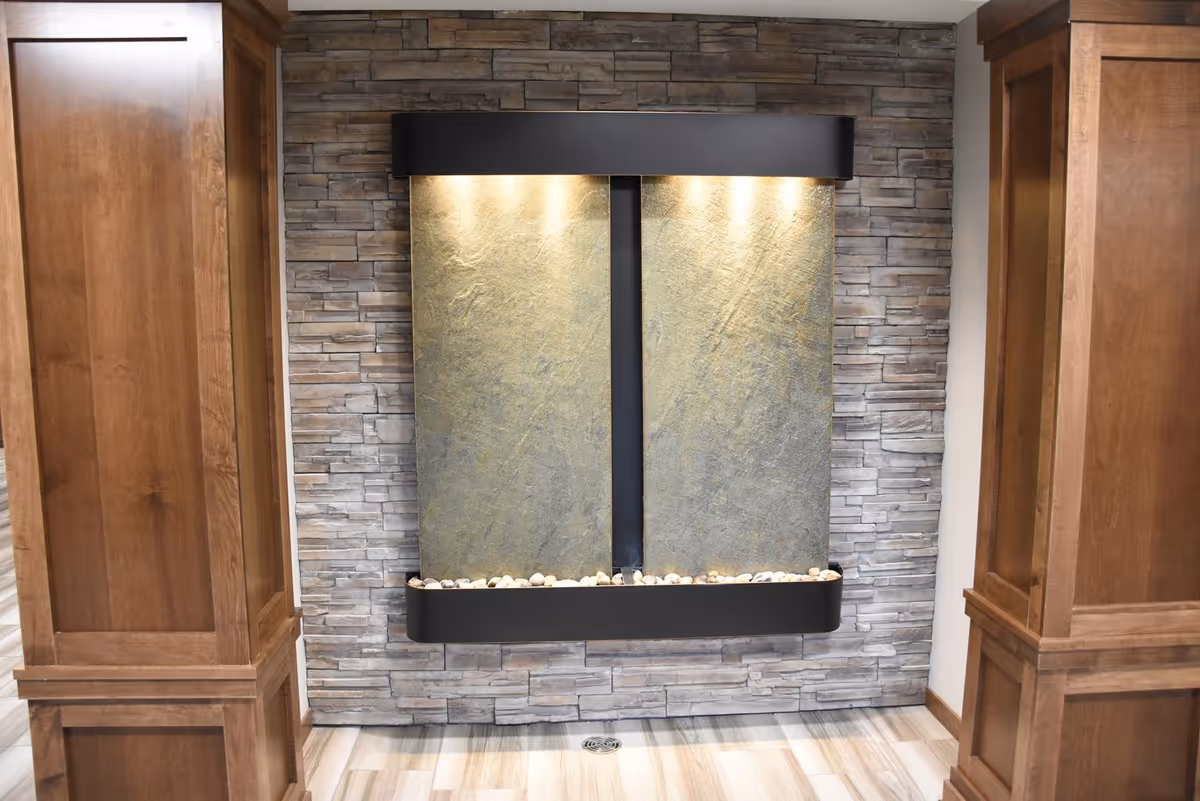 Indoor water feature with two vertical stone panels illuminated by overhead lights, set against a wall with gray stone tiles, flanked by two wooden pillars on either side and a floor with light-colored wood-like tiles.