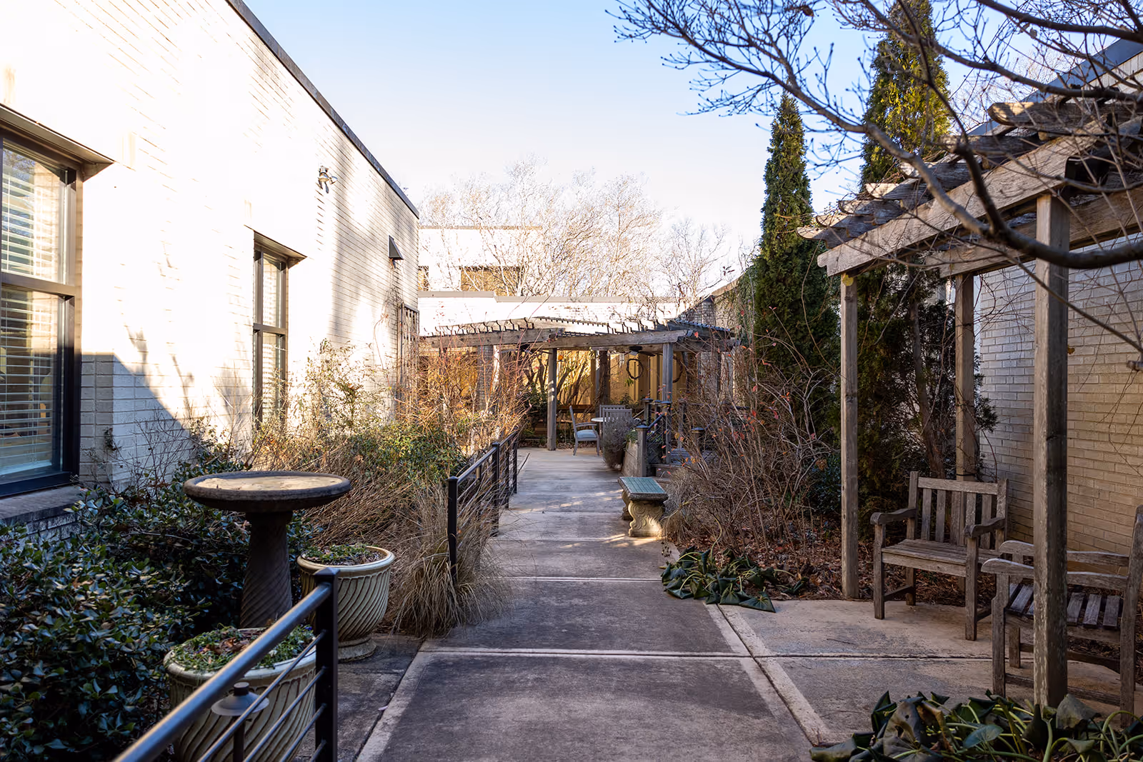 Outdoor walkway between buildings at A.G. Rhodes Atlanta with benches, potted plants, a birdbath, and a wooden pergola. Leafless trees and shrubs line the path under a clear sky.