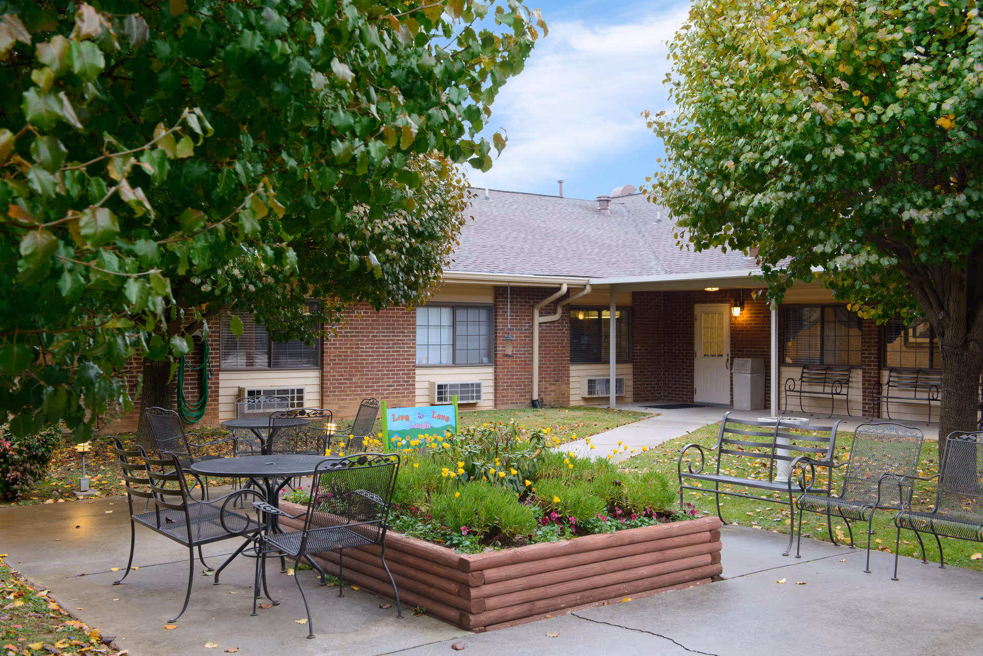 Outdoor courtyard with metal tables and chairs, a raised flower planter, and trees in front of a brick senior living building.