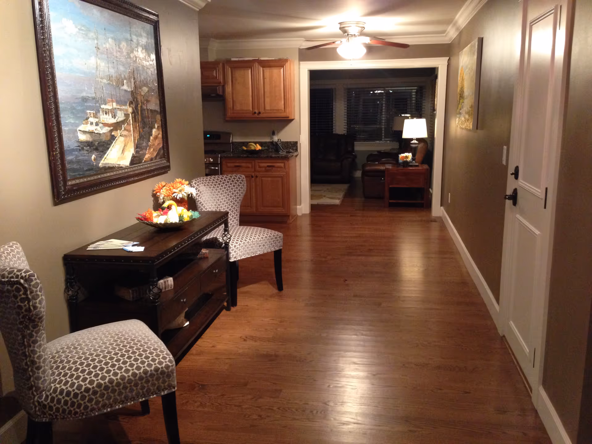 A well-lit interior hallway with hardwood floors, patterned chairs and a console table leading to a small kitchen and living room.