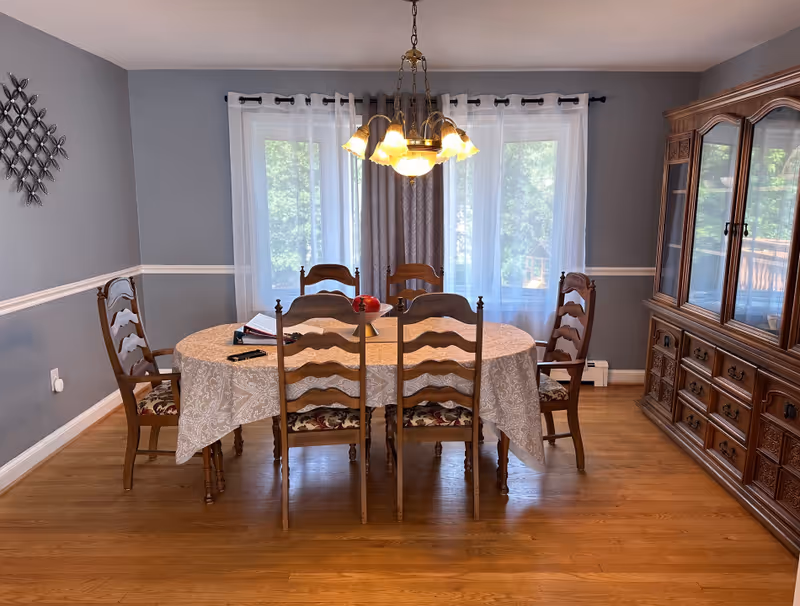A dining room with a wooden table covered by a beige patterned tablecloth and six wooden chairs with floral cushions. A chandelier with multiple lights hangs above the table. Behind the table are two large windows with sheer white curtains and gray drapes. To the right is a large wooden china cabinet with glass doors and drawers. The walls are painted gray with white trim, and the floor is hardwood.