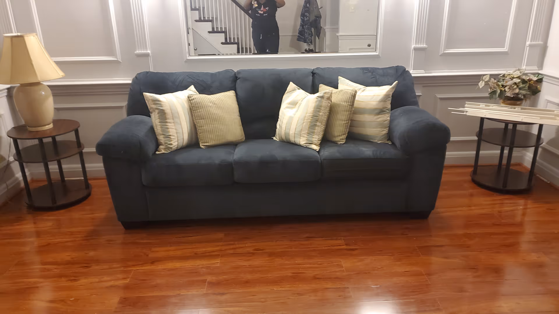 A dark blue sofa with five striped and solid-colored cushions is placed against a white paneled wall with a large mirror above it. On either side of the sofa are round wooden side tables; the left table has a beige lamp, and the right table has a floral arrangement. The floor is polished wood.