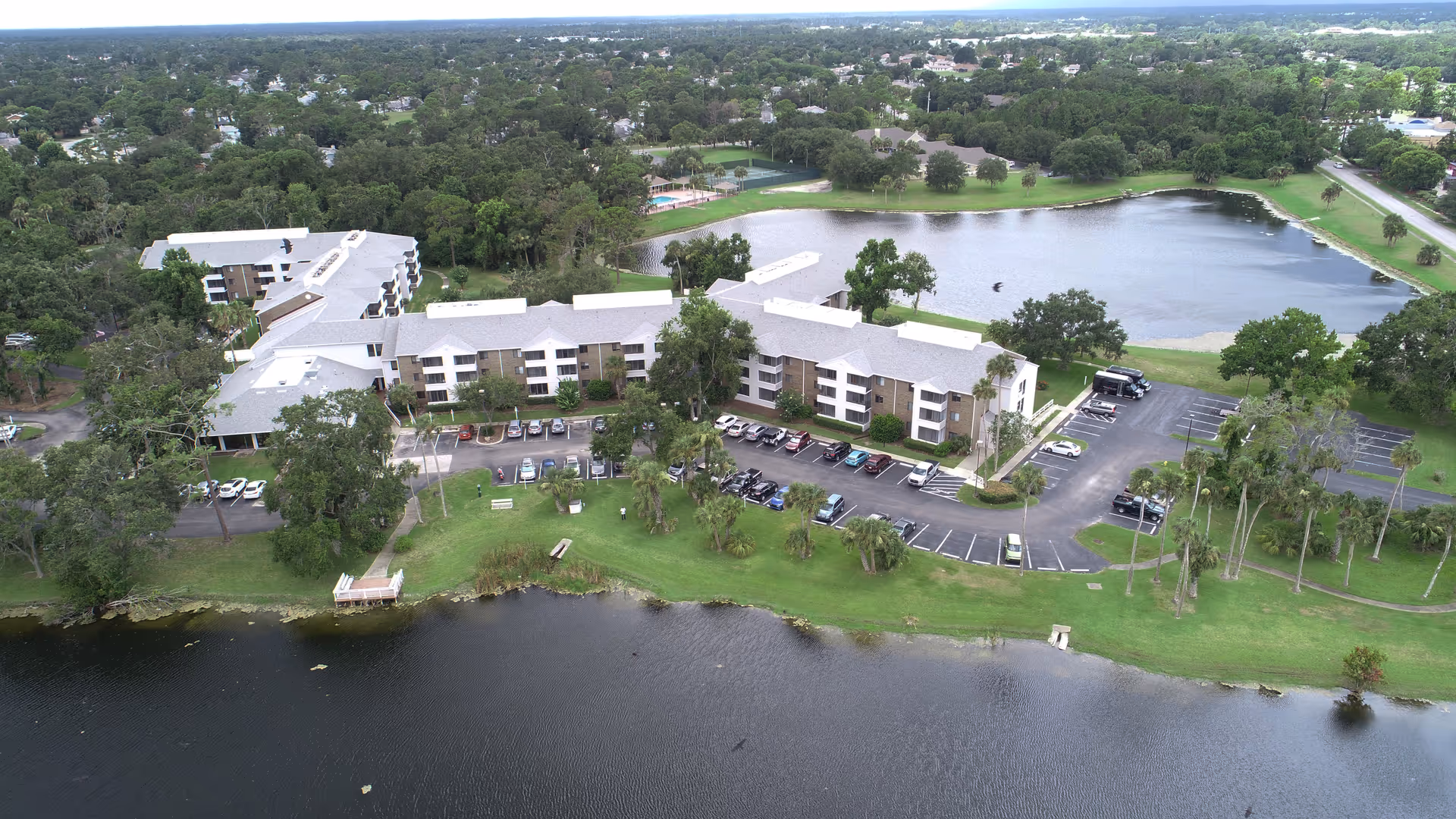 Aerial view of a senior living facility named CountrySide Lakes, showing multiple connected buildings with white roofs surrounded by trees and greenery. There is a large pond or lake in the foreground and another body of water behind the buildings. Several cars are parked in parking lots near the buildings, and there are grassy areas with benches and walking paths.