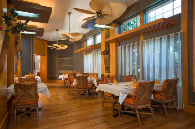 A dining room with several tables covered in white tablecloths, each set with glasses, napkins, and small flower arrangements. The room features wooden floors, large windows with sheer curtains, ceiling fans with leaf-shaped blades, and warm lighting.