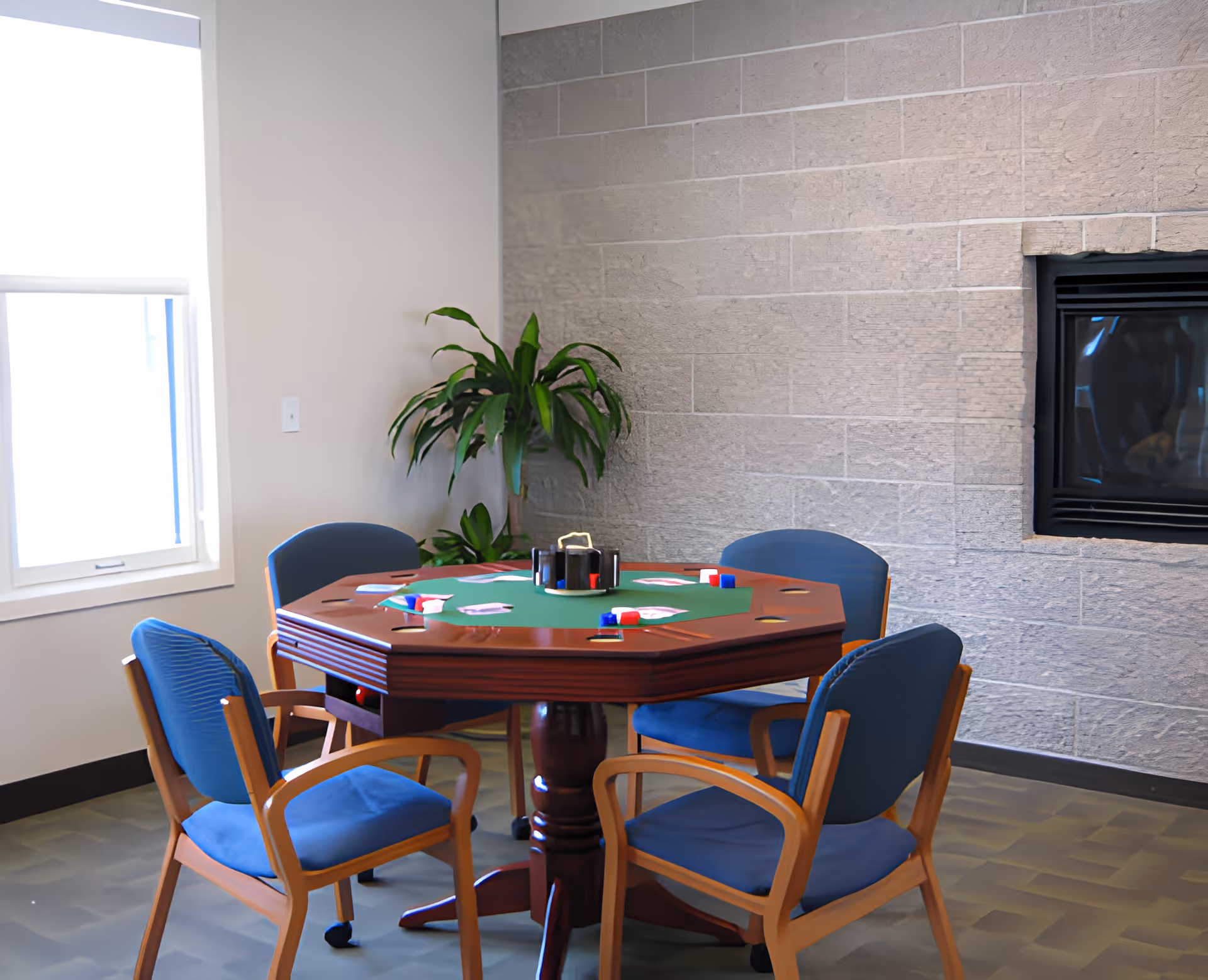 An octagonal card table with poker chips surrounded by four blue upholstered chairs in a bright room with a potted plant and a wall-mounted fireplace.