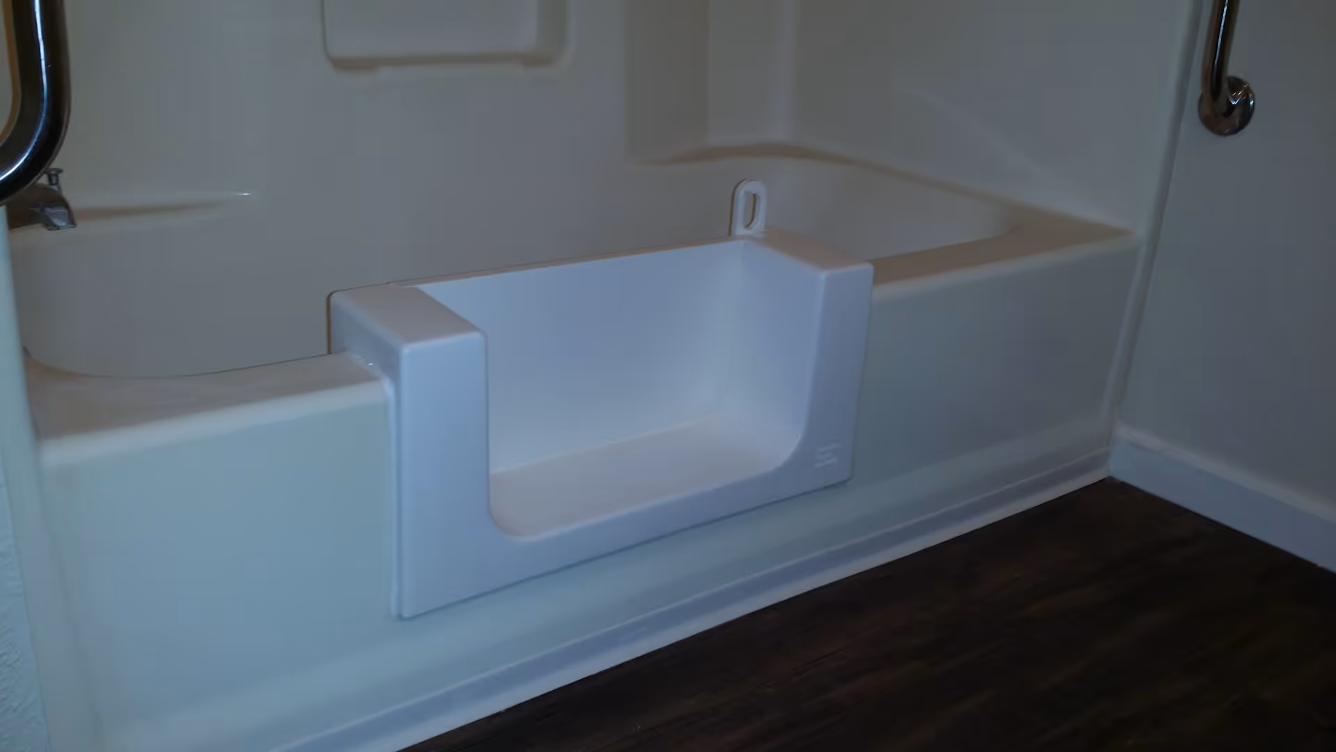 A bathtub with a built-in seat and a cut-out section for easy access, featuring a grab bar on the wall for safety, and dark wood flooring outside the tub area.