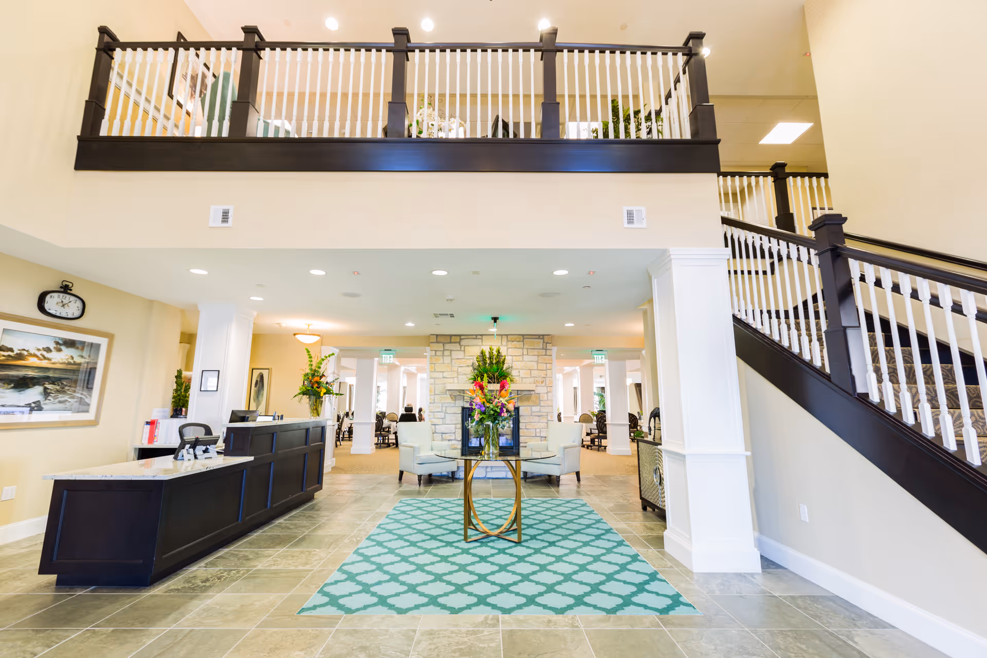 Bright two-story lobby and reception area with a front desk, staircase, seating, and a floral centerpiece on a patterned rug.