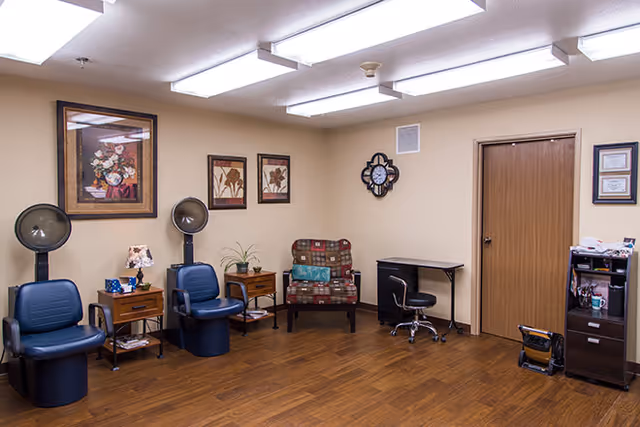 Interior room with two blue salon hair dryer chairs, a patterned armchair, small wooden side tables, a desk with a rolling chair, and framed artwork on beige walls. The floor is wood and the ceiling has fluorescent lights.