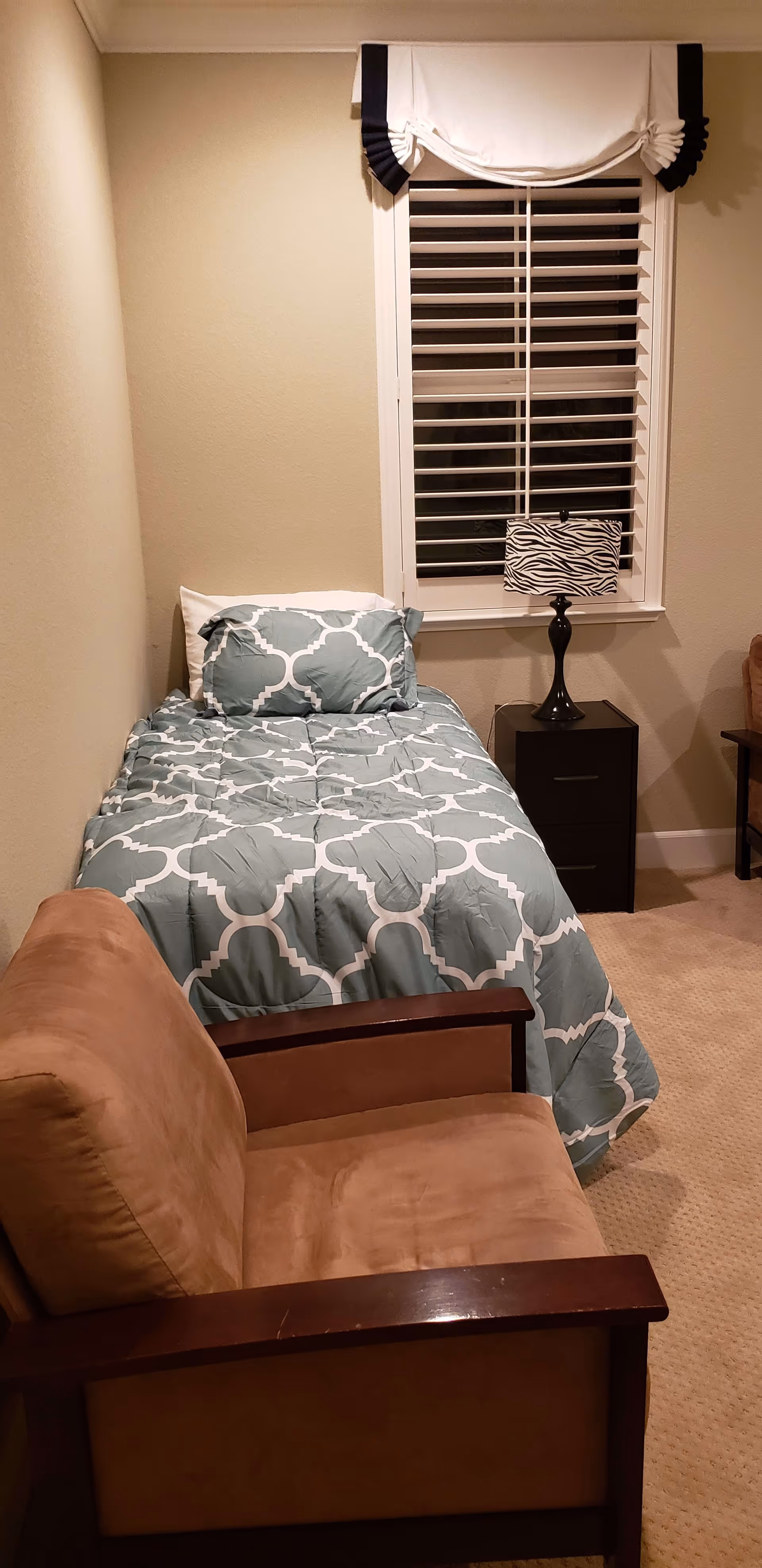 A small bedroom with a single bed covered with a blue and white patterned comforter and matching pillow. Next to the bed is a black nightstand with a lamp that has a zebra print lampshade. In the foreground, there is a brown cushioned armchair with wooden armrests. The room has beige walls and carpet, and a window with white blinds and a white and black valance above it.