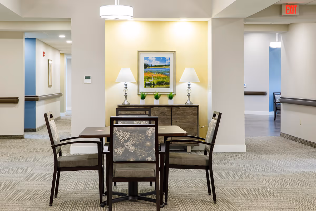 A small seating area in a hallway of an assisted living facility with a square wooden table and four cushioned chairs with floral patterned backs. Behind the table is a wooden sideboard with two table lamps and three small potted plants. Above the sideboard hangs a framed landscape painting. The walls are light-colored with a yellow accent wall behind the sideboard. The floor is carpeted, and there are hallways visible on either side with handrails and an exit sign in the background.
