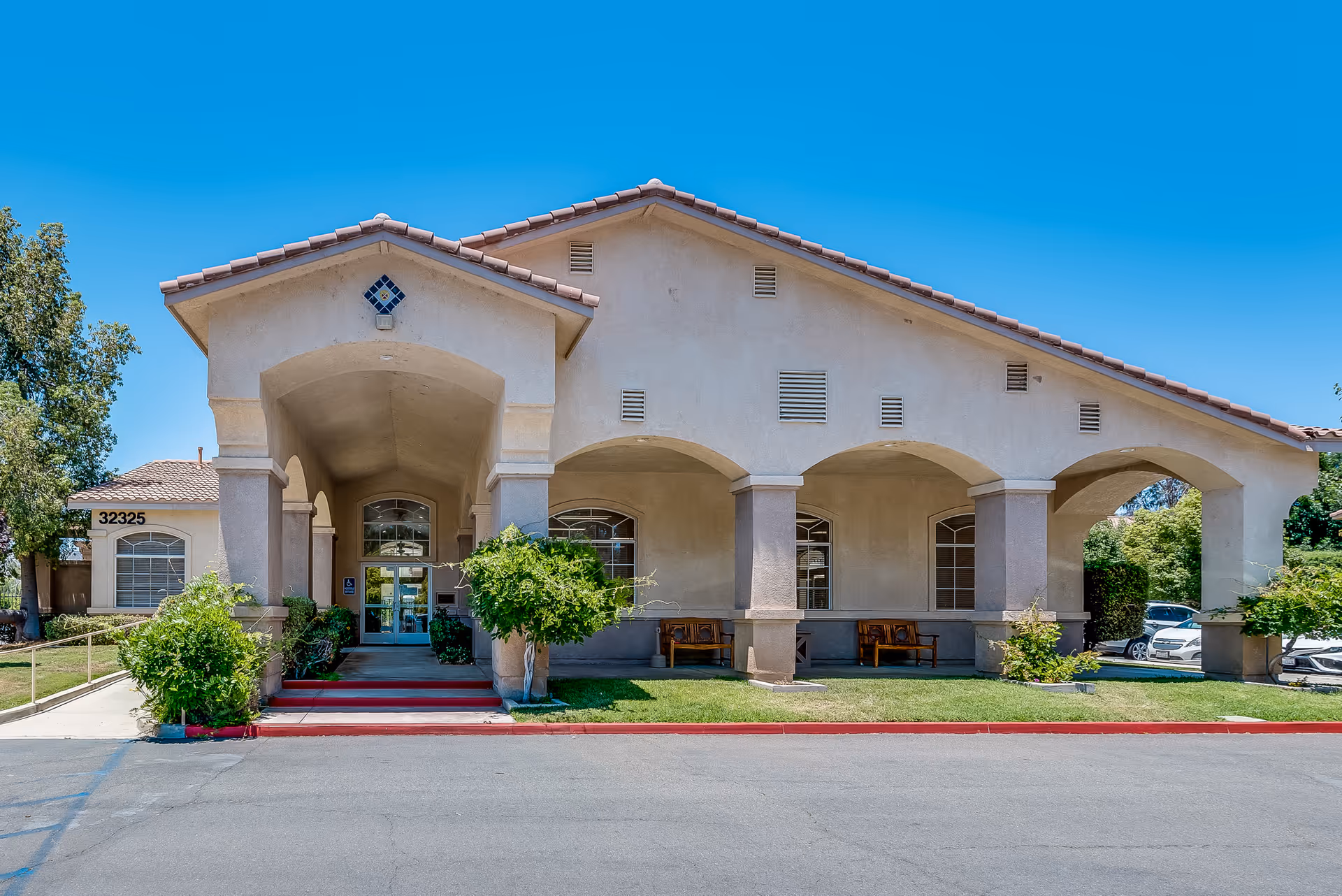 Front exterior view of Wildomar Senior Assisted Living facility with a beige stucco building, arched entrance, tiled roof, and a few benches under the covered porch. There are some green bushes and trees around the entrance and a clear blue sky above.