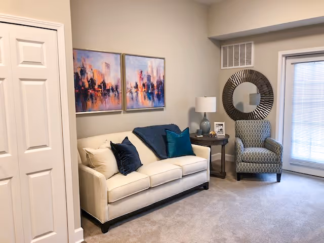 A cozy living room area featuring a cream-colored sofa with multiple throw pillows in shades of blue and beige. Above the sofa are two abstract cityscape paintings with vibrant colors. To the right of the sofa is a round wooden side table with a lamp, a framed photo, and decorative items. Next to the table is a patterned armchair in gray and white, positioned near a glass door with blinds. A round decorative mirror hangs on the wall above the armchair. The room has beige carpet and neutral-colored walls.