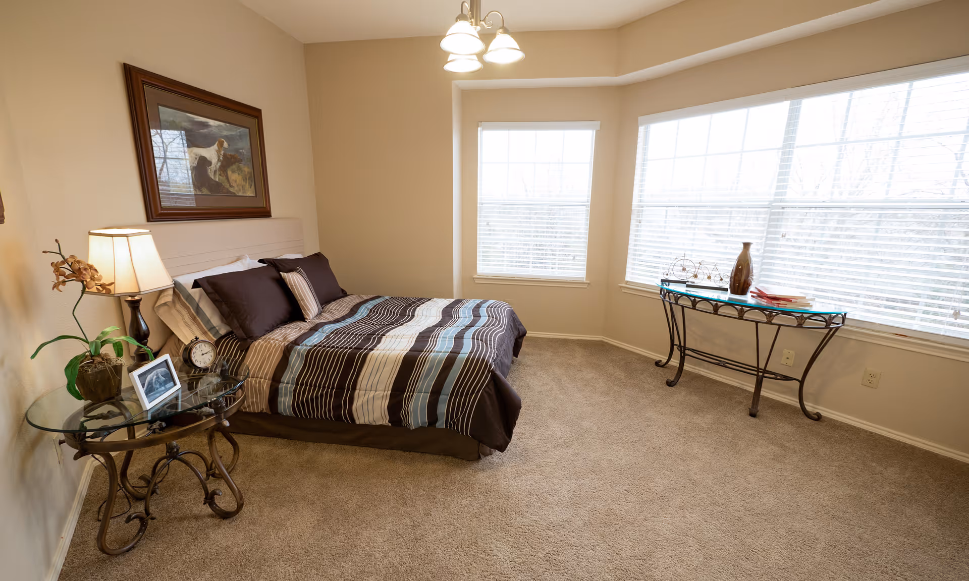A bedroom with a bed covered in a striped brown, beige, and blue comforter. There is a glass-top side table with a lamp, a plant, a clock, and a framed photo next to the bed. A framed picture of dogs hangs on the wall above the bed. The room has beige walls, carpeted floor, and large windows with white blinds letting in natural light. A glass-top console table with decorative items is placed under the windows.