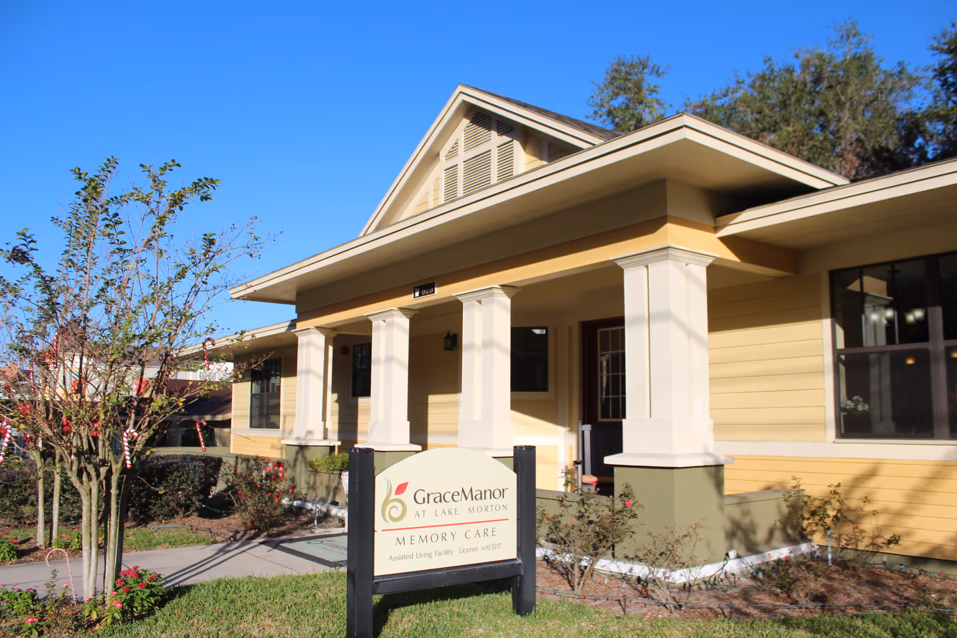 Exterior view of Grace Manor at Lake Morton, a single-story yellow building with white columns and a covered porch, surrounded by landscaping and a clear blue sky.