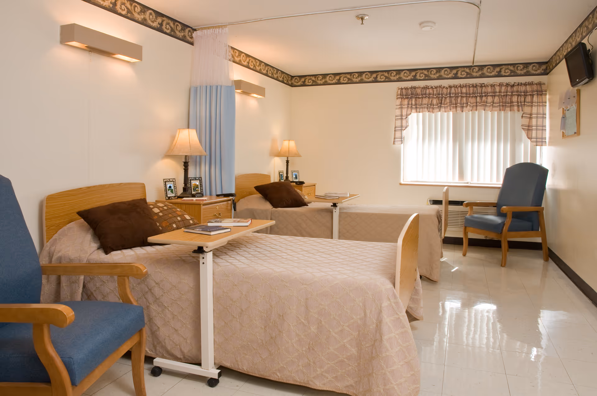 A clean and well-lit room with two single beds, each with a beige quilted bedspread and brown pillows. Each bed has an overbed table with books on it. Between the beds is a nightstand with two lamps and framed photos. There are two blue armchairs, one near the foot of each bed. The room has a window with vertical blinds and a plaid valance, and a small wall-mounted TV in the corner.
