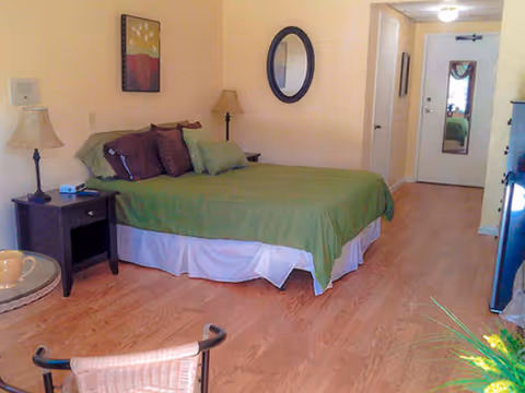 A bedroom with a bed covered in green bedding and multiple pillows. There is a nightstand with a lamp and an alarm clock next to the bed. A round mirror hangs on the wall above the bed, and a framed picture is on the adjacent wall. The room has wooden flooring, a chair in the foreground, and a door with a mirror on it at the far end.