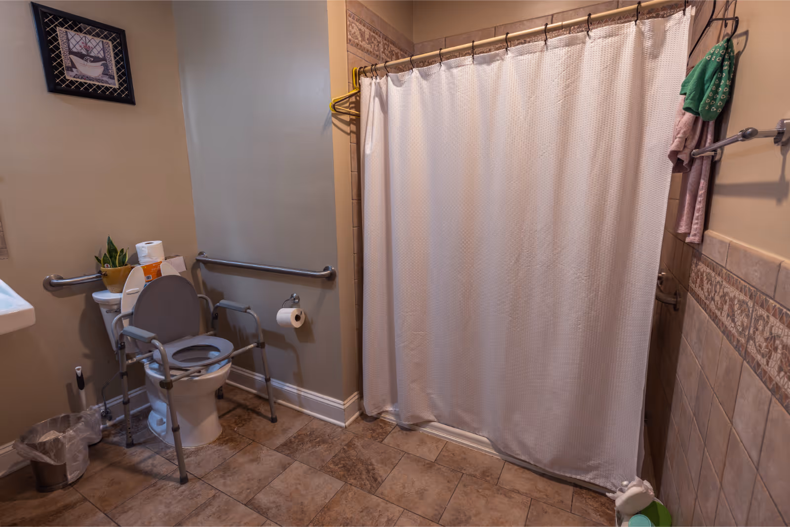 A bathroom with a toilet equipped with safety rails and a raised seat, a white shower curtain, a wall-mounted grab bar, a small plant on the toilet tank, a trash can with a liner, and towels hanging on a rack.