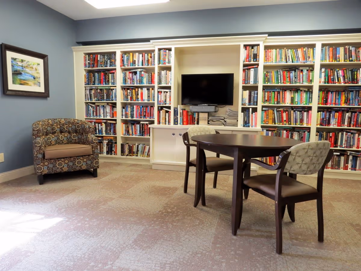 A cozy room with a patterned armchair on the left, a round wooden table with two chairs in the center, and a large white bookshelf filled with books along the back wall. A flat-screen TV is mounted in the middle of the bookshelf, and a framed picture hangs on the blue wall to the left.