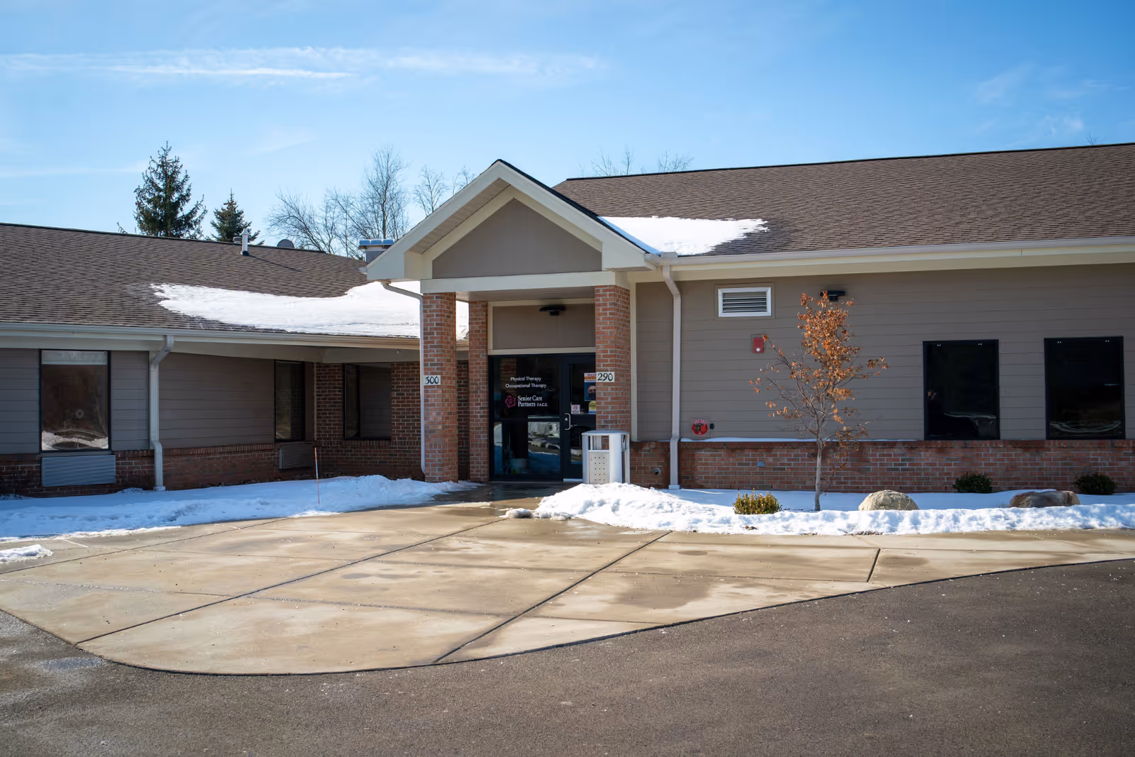Entrance of a single-story senior care facility with a covered portico, snow on the ground and roof.