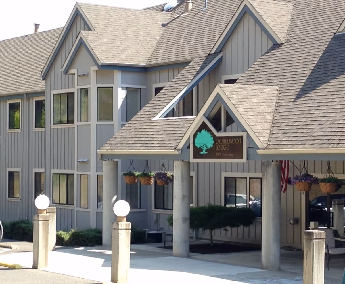 Exterior view of a senior living facility building with gray siding and multiple windows. The entrance has a covered porch supported by concrete pillars, hanging flower baskets, and a sign that reads 'Laurelwood Lodge'. There are round white light fixtures on short concrete posts near the entrance.