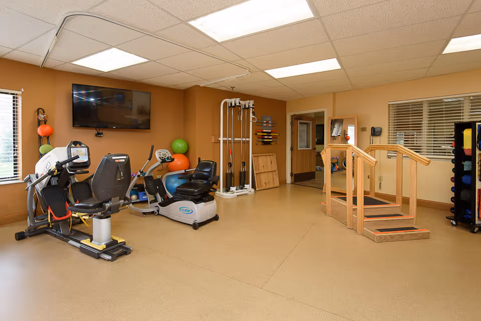 A well-lit exercise room with various fitness equipment including a recumbent bike, elliptical machine, exercise balls, resistance bands, and a wooden staircase for physical therapy. A large flat-screen TV is mounted on the wall, and there are windows with blinds letting in natural light.