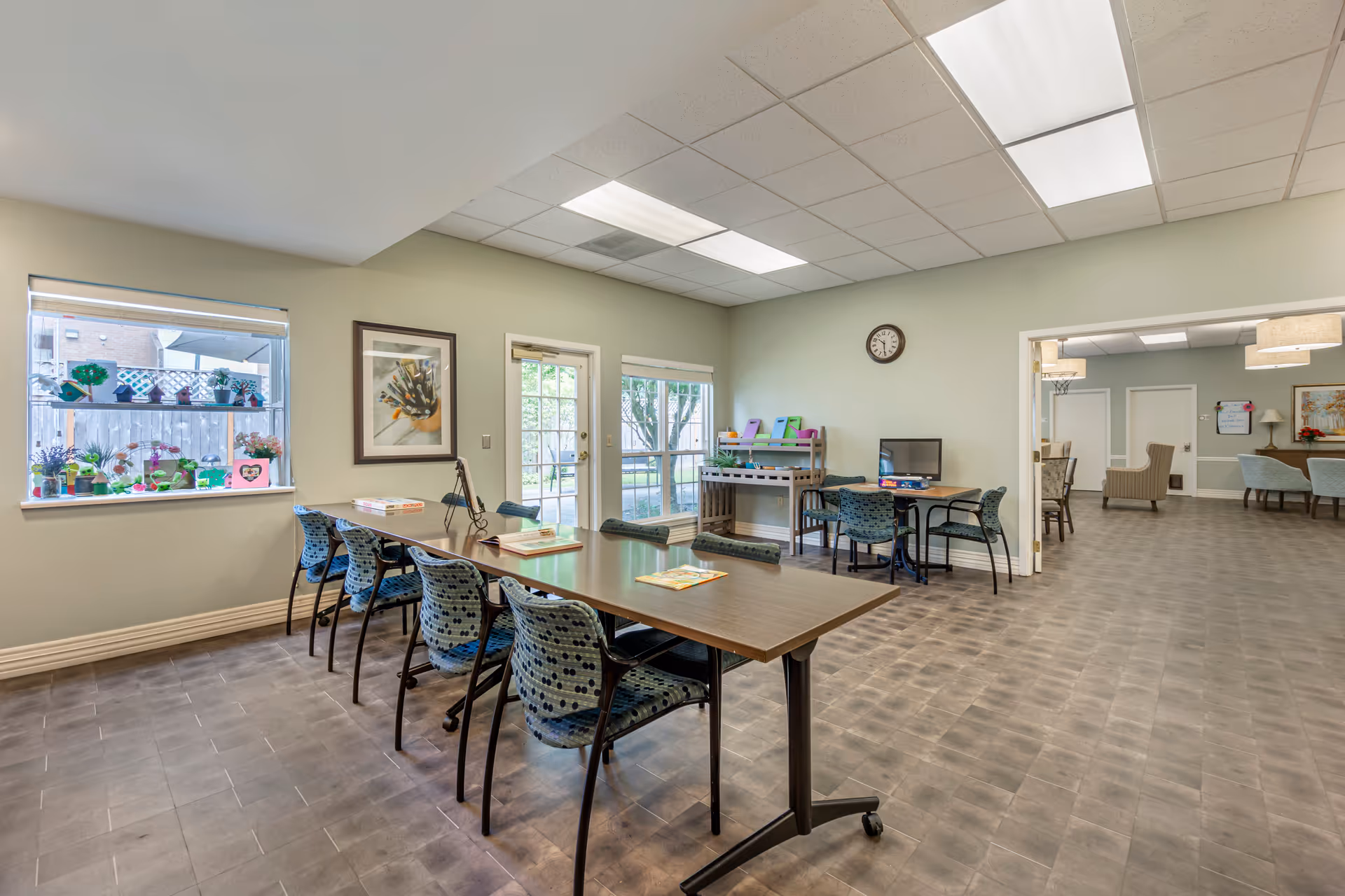 A bright activity room in a senior living facility with a long table surrounded by blue patterned chairs. The room has large windows and a glass door leading outside. There are colorful crafts displayed on the window sill and a small table with a computer and board games. In the background, there is an adjoining room with more seating and a basketball hoop on the wall.