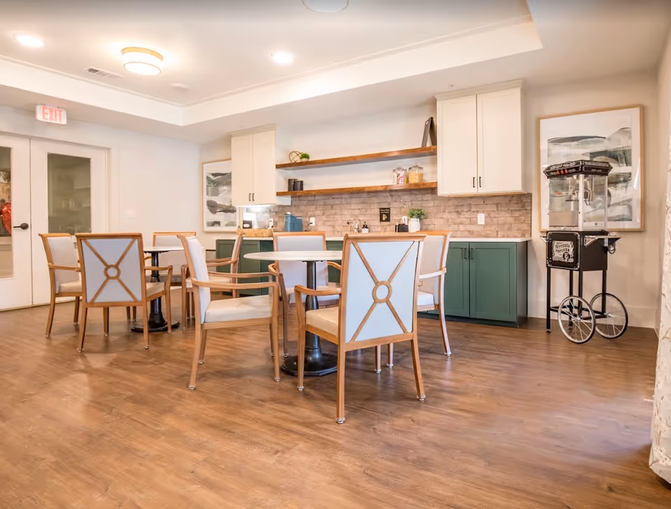Bright communal dining area with round tables and upholstered chairs in front of a kitchenette with green lower cabinets and open shelves.