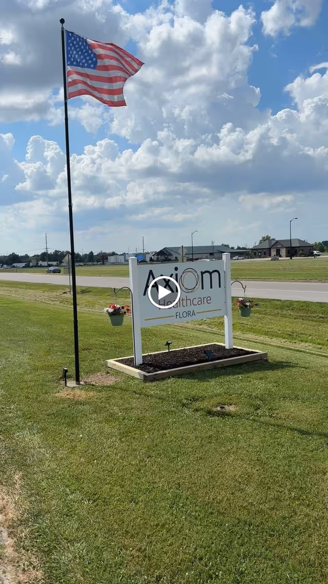 A white sign for Axiom Healthcare Flora on a grassy lawn beside a road with an American flag on a pole and buildings in the background.