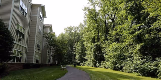 Paved path alongside a multi-story apartment building bordered by lawn and dense trees.