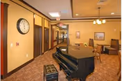 Communal interior with a grand piano, booth seating, tables, and a wall clock in a retirement center.