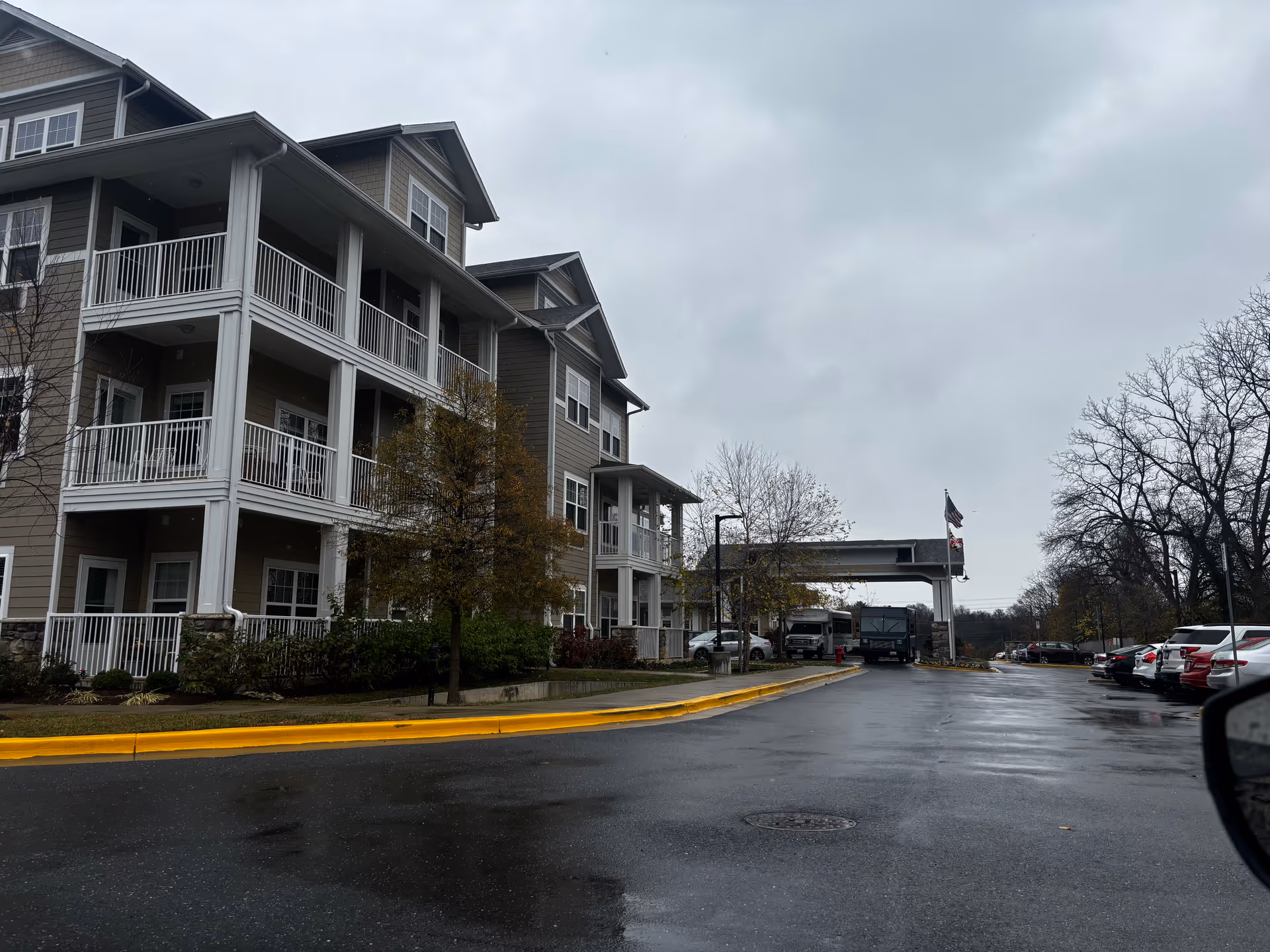 Gray three-story senior living building with white balconies, a covered drive-up entrance, and a wet parking area on an overcast day.