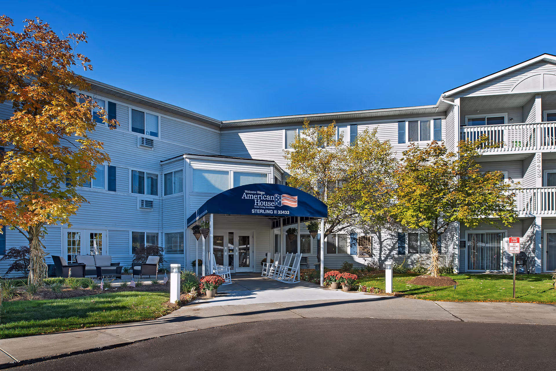 Front entrance of the American House Sterling Meadows senior living building with a blue covered awning, rocking chairs, and landscaped trees.