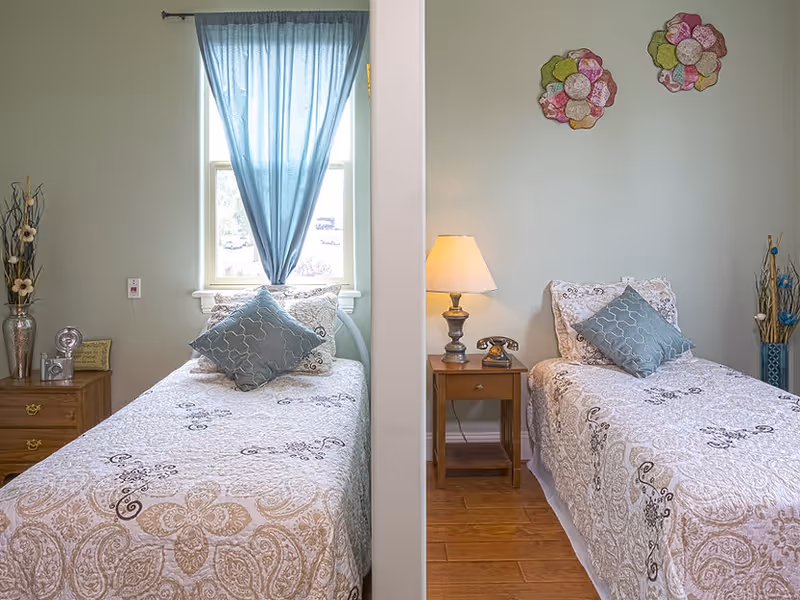 Two twin beds in a pastel-themed shared bedroom with a central divider, bedside tables, a lamp, and decorative pillows.