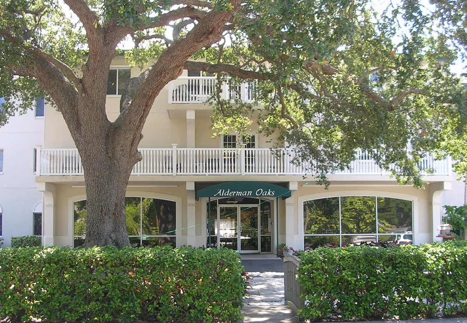 Front exterior view of Alderman Oaks Retirement Residence showing a large tree and green bushes in front of the building entrance with a green awning displaying the facility name.